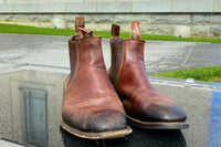 Before and after R.M. Williams boots resoled by Shane's Shoe Repairs, photographed outside the Auckland Museum showing restored leather and new soles.
